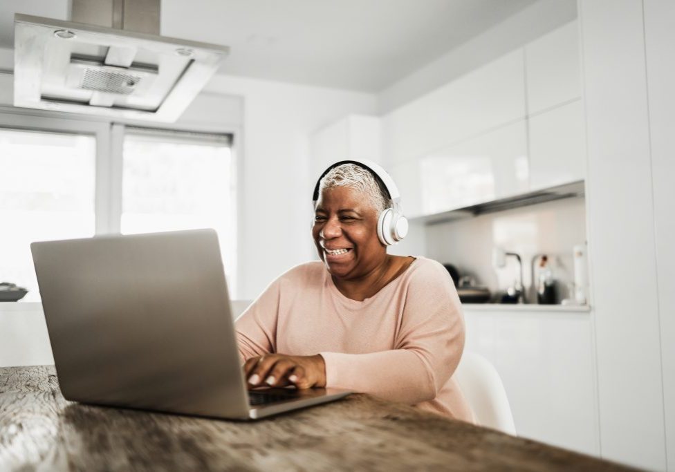 Senior african woman using laptop computer while wearing headphones at home - Focus on face